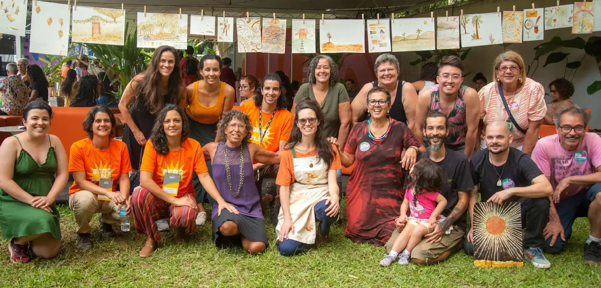 Grupo de participantes da oficina Aquaterra Rupestre na FLIP, com as aquarelas de terra penduradas em varal ao fundo