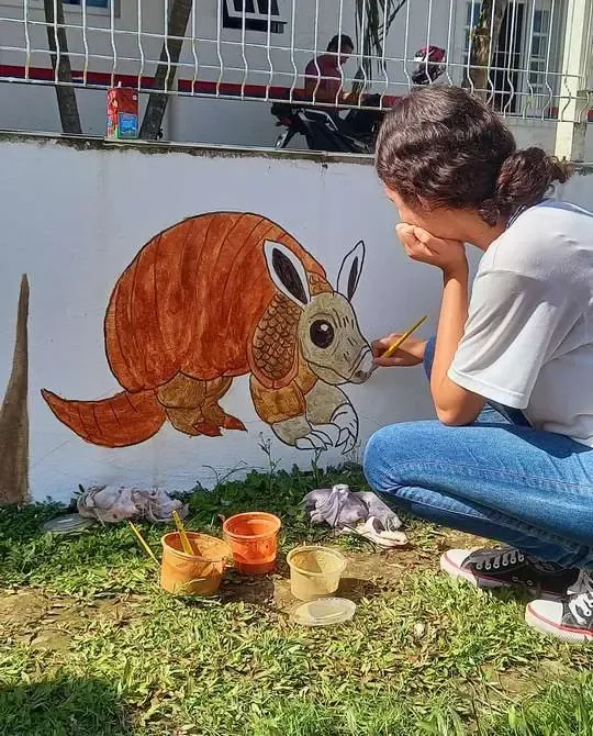 Estudante pintando um tatu em tinta de terra no muro da escola, com potes de pigmento no chão