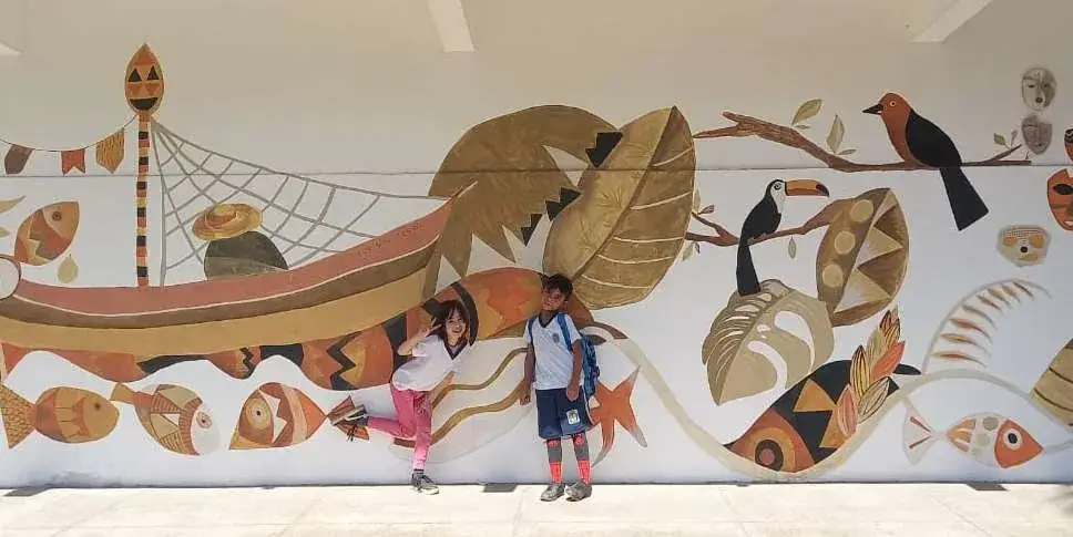 Mural em tintas de terra com canoa, peixes, tucano e folhas em tons de ocre e terracota, com duas crianças em frente