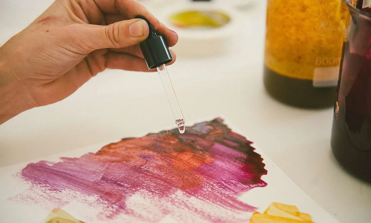 Pigmento líquido sendo pingado com conta-gotas sobre pintura em tons de rosa e vinho
